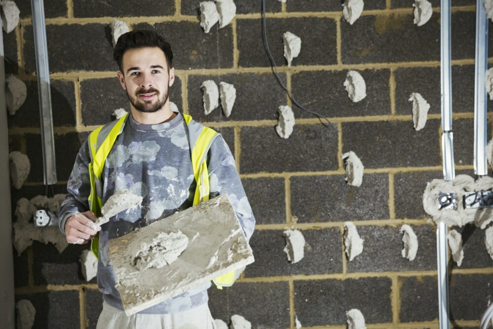 A plasterer, a builder with plasterboard and spreading trowel in front of a cinder block wall.