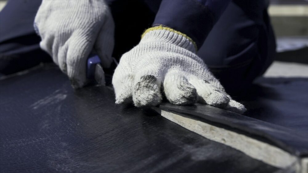 Close up for worker hands in white protective gloves cutting a black sheet of ruberoid with a